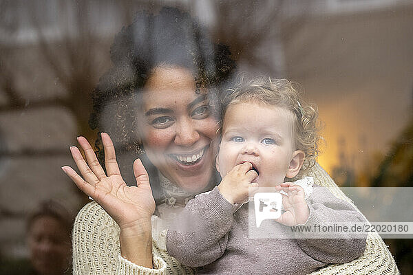 Woman smiling and waving with a baby girl through a glass window indoors. Netherlands