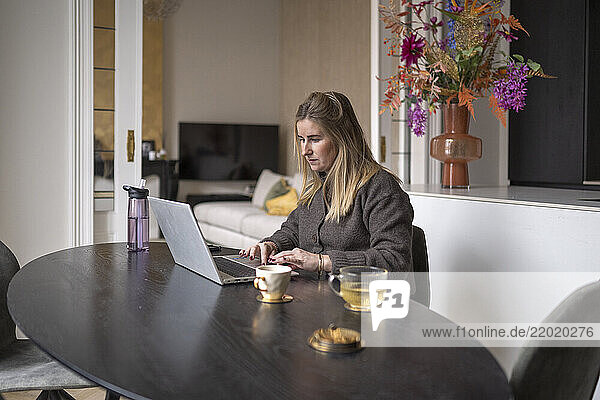 A woman working at a laptop at a dark wooden table in a cozy living space  surrounded by colorful flowers. Netherlands