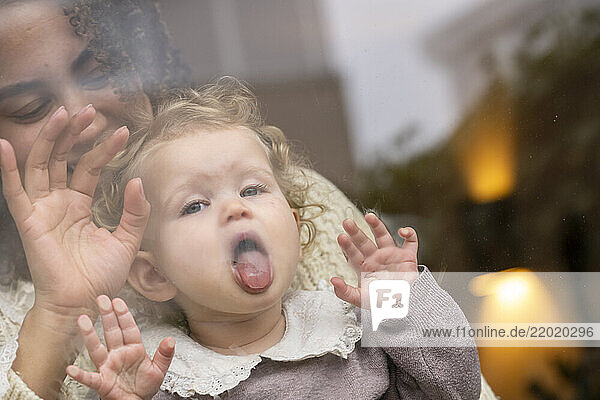 Baby with curly hair pressing face against glass  mother in background smiling and waving. Netherlands