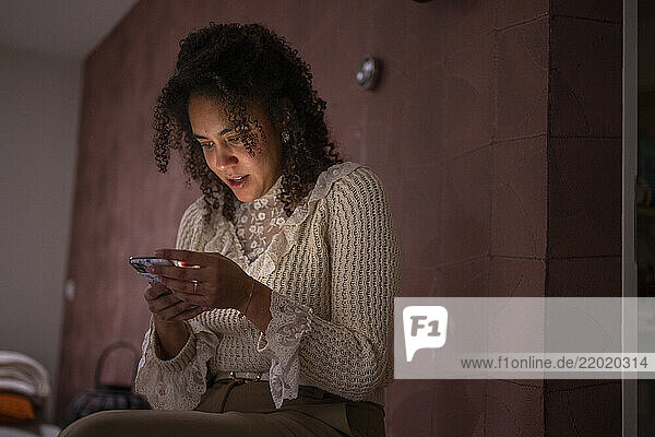 Woman with curly hair in a sweater using a smartphone  smiling in a warmly lit room. Netherlands