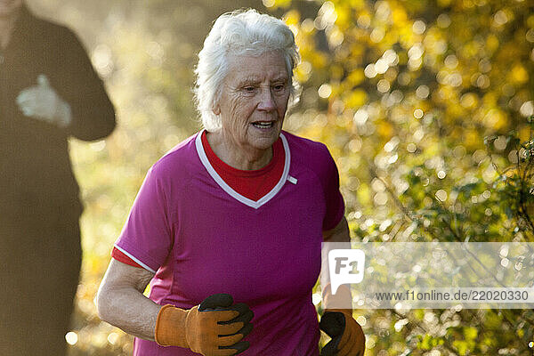 An elderly woman with white hair jogs energetically in a park  wearing a bright pink shirt and orange gloves. Sale Water Park  Manchester  UK