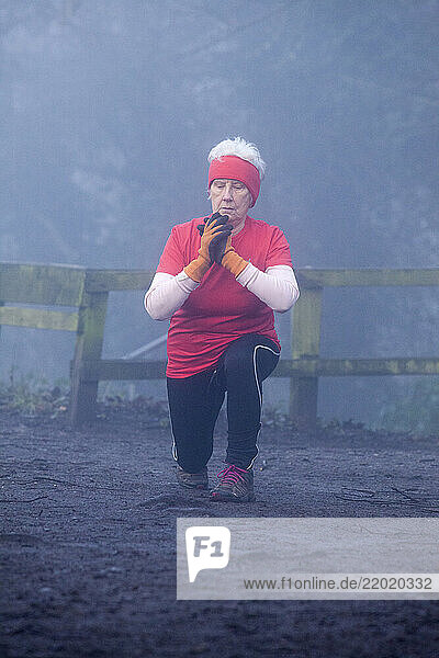 Elderly woman in sportswear doing lunges outdoors in a foggy forest  wearing a red headband and shirt  gloves  and black leggings. Sale Water Park  Manchester  UK