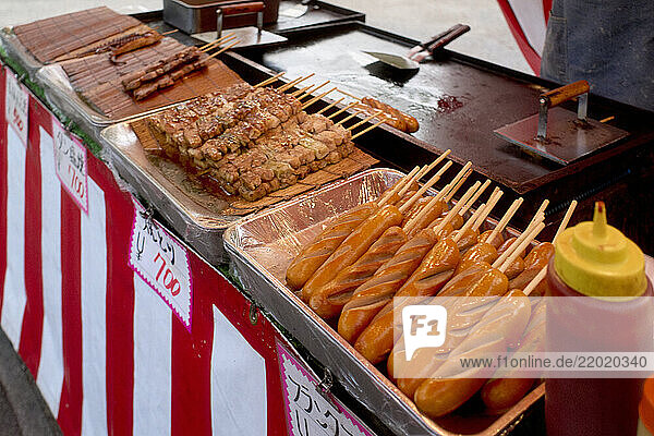 Assorted grilled skewers and corn dogs on a street food vendor s striped counter. Osaka  Japan