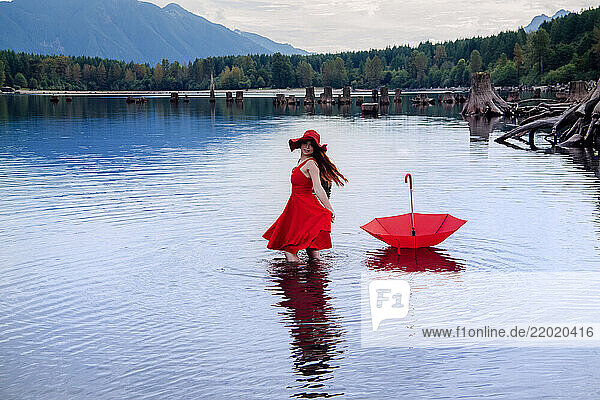 Woman in red dress and hat stands in lake near a red umbrella  surrounded by mountains and trees. Rattlesnake Lake  Puget Sound  WA  USA