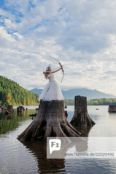 Woman in a white dress with a bow and arrow standing on a large tree stump in a lake. Rattlesnake Lake  Puget Sound  WA  USA