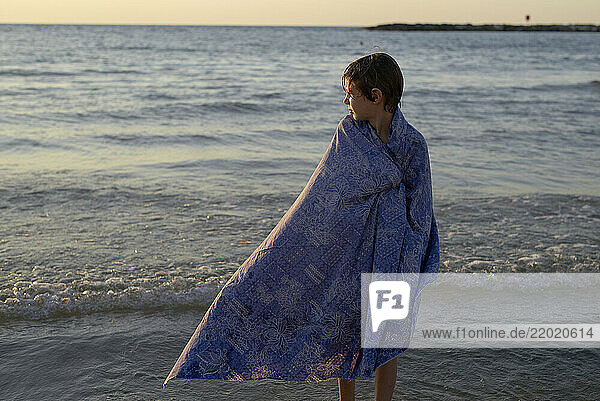 Child wrapped in a blue towel standing by the sea at sunset. Israel