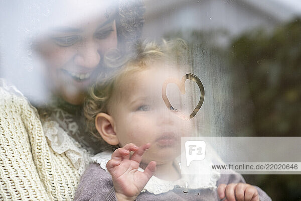 Smiling woman holds baby drawing a heart on a foggy window inside a cozy setting. Netherlands