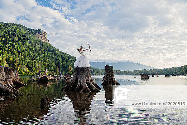 Woman in a white dress aiming a bow from a tree stump in a serene lake landscape. Rattlesnake Lake  Puget Sound  WA  USA