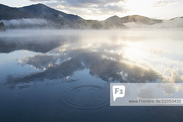 Morning mist over calm Lake Placid surface reflecting clouds and sunlight