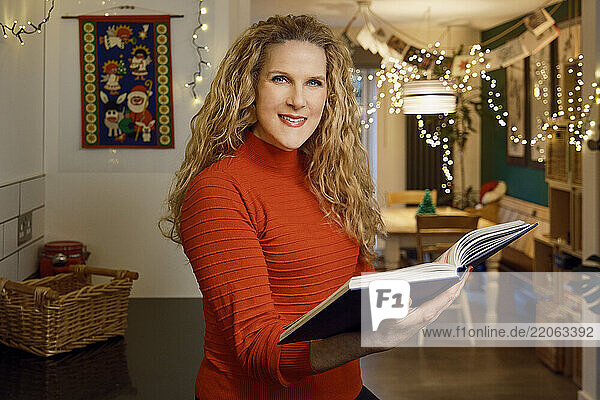 Happy mature woman holding cookbook in Christmas at home