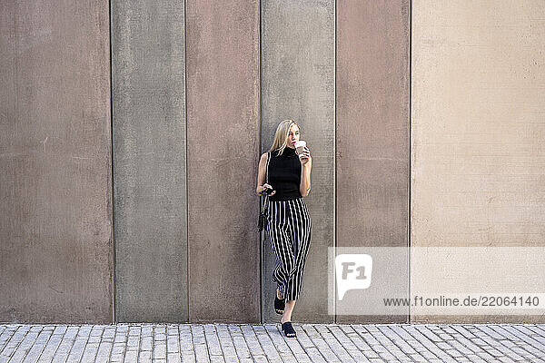 Young blond woman with cell phone leaning against wall drinking coffee to go