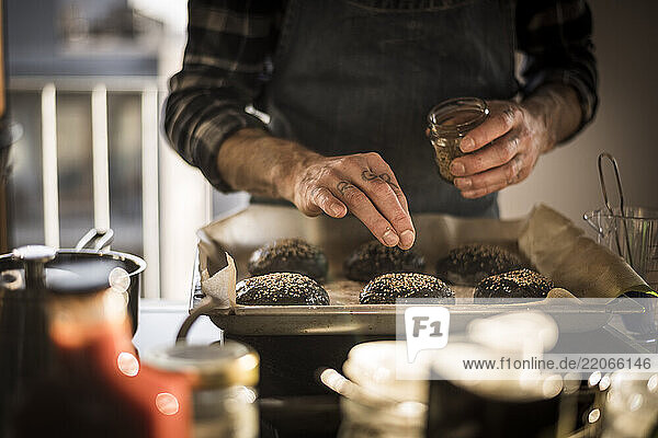 Man sprinkling sesame on burger buns on a baking tray