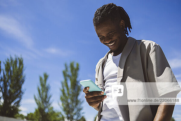 Happy young man using smart phone under blue sky in park
