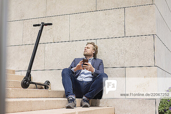 Businessman with E-Scooter and smartphone sitting on steps relaxing