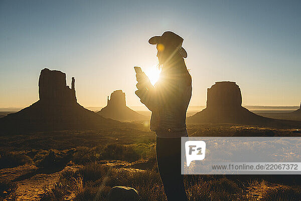 USA  Utah  Monument Valley  silhouette of woman with cowboy hat using mobile phone at sunrise