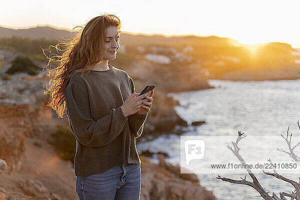 Redheaded young woman using cell phone at the coast at sunset  Ibiza  Spain