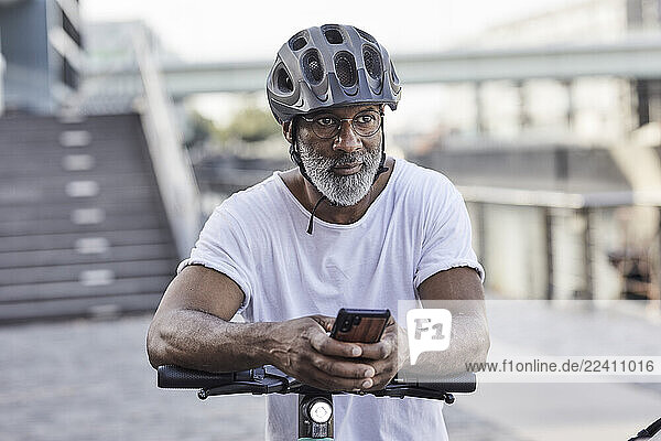 Portrait of pensive mature man wearing cycling helmet leaning on handlebar of Electric Scooter