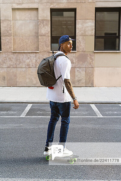 Tattooed young man with backpack standing on skateboard looking around