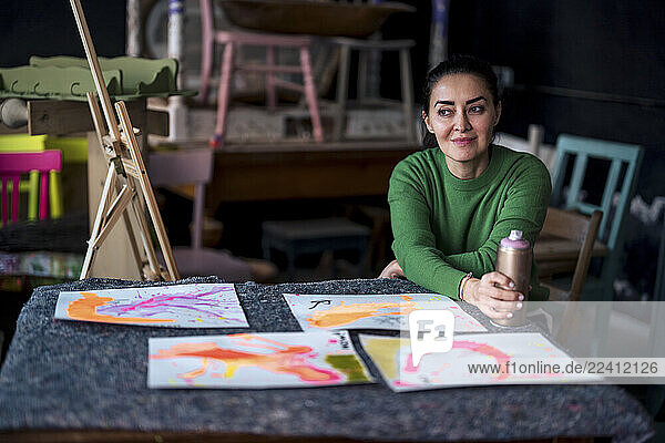 Female artist in green sweater smiling in art studio with paintings and spray can