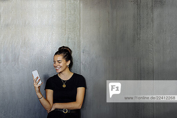 Portrait of happy casual young businesswoman using smartphone in front of a grey wall