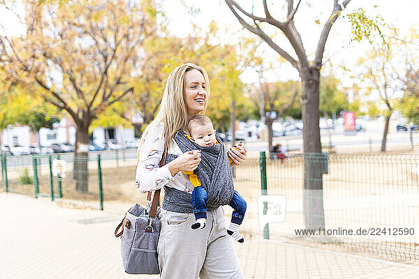 Happy mother walking with baby boy in sling outdoors