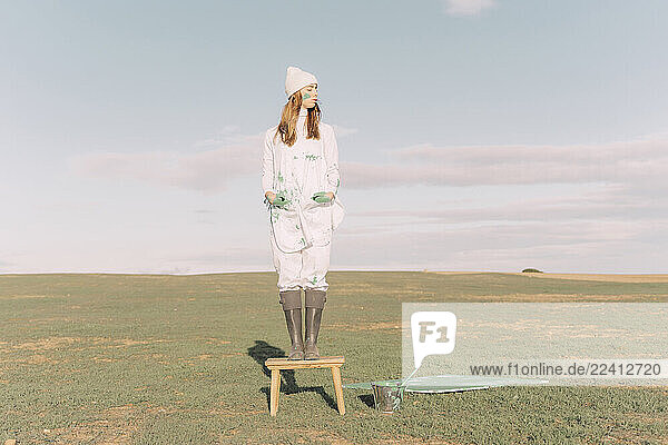 Young woman standing on stool by green painting on dry field