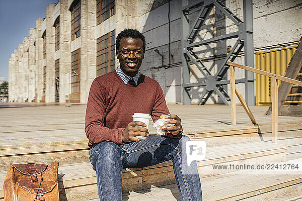 Young man sitting on stairs in the city  eating hamburger  drinking coffee