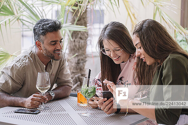 Happy friends sitting at table with drinks looking at smartphone