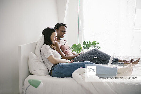 Two happy women lying in bed at home looking at laptop