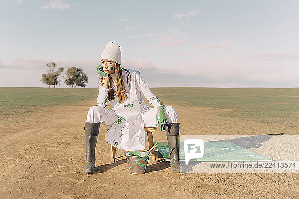 Young woman sitting on stool with green painting on dry field