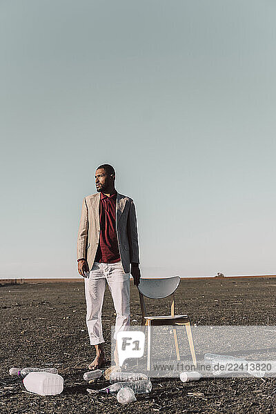 Young man standing next to chair surrounded by plastic bottles in barren land