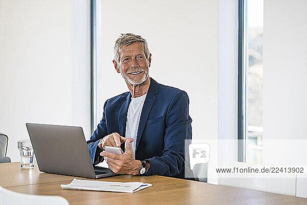 Smiling senior businessman with laptop and cell phone sitting at desk at home