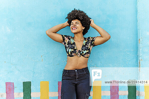 Smiling Afro-American woman in front of a blue wall with hands in hair