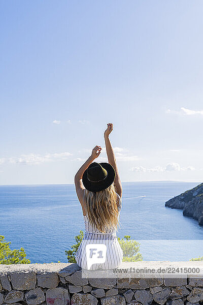 Young woman sitting on a wall with raised arms and looking at distance  viewpoint in Ibiza