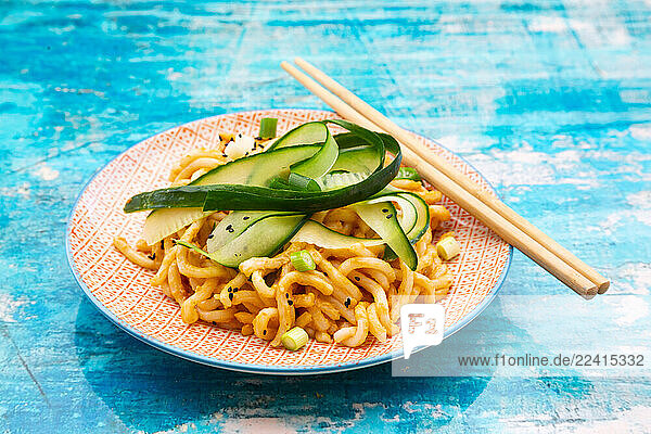 Udon noodles with peanut butter sauce and cucumber on a plate with chopsticks on a blue background