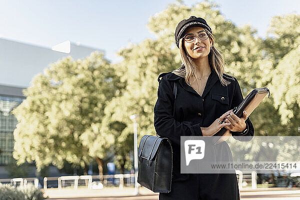 Young blond businesswoman wearing black sailor s cap and holding laptop bag and looking sideways