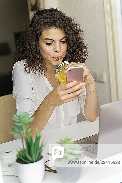 Young woman using cell phone and drinking orange juice at desk at home