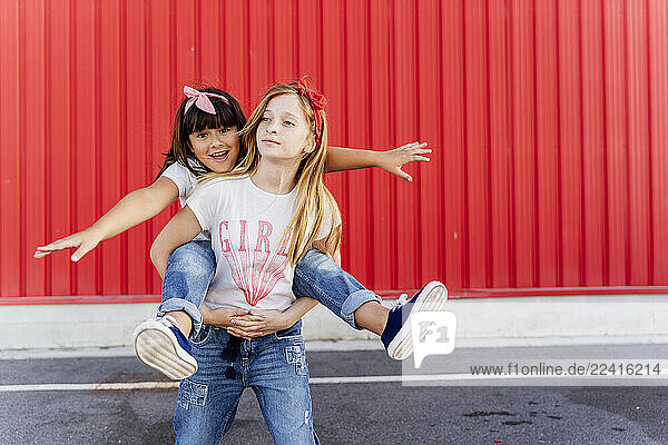 Girl carrying her sister on back  standing in front of a red wall