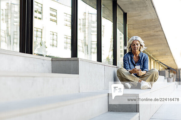 Mature businesswoman sitting cross-legged on steps in the city  using smartphone