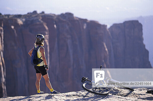 A female mountain biker stands on the edge of a cliff and looks out at the horizon.
