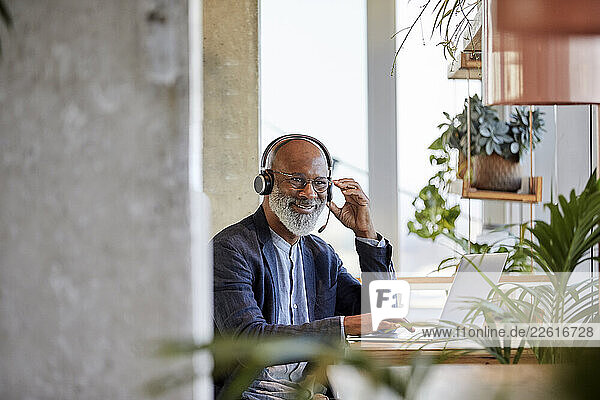 Smiling businessman listening through headphones while sitting with laptop at table and working from home