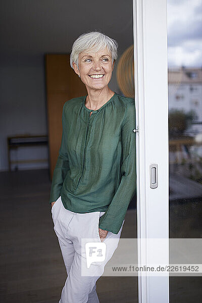Smiling woman with white hair in a green blouse leaning against a glass door at home