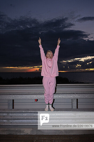 Young woman with pink hair wearing pink track suit shirt standing on road barrier at night