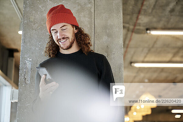 Man wearing red hat text messaging on smart phone standing against column at home