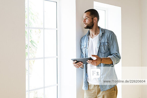 Young serious man at home holding tablet standing at window and looking outside