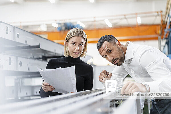 Businessman and woman with papers examining metal rods in factory hall
