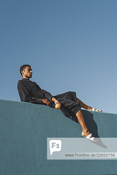Young man wearing black kaftan lying on blue wall holding oranges