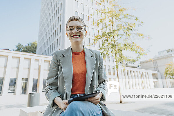 Mid adult woman smiling while holding digital tablet sitting on street during sunny day