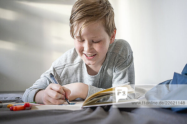 Portrait of smiling boy lying on bed using felt tip pens and sketch book
