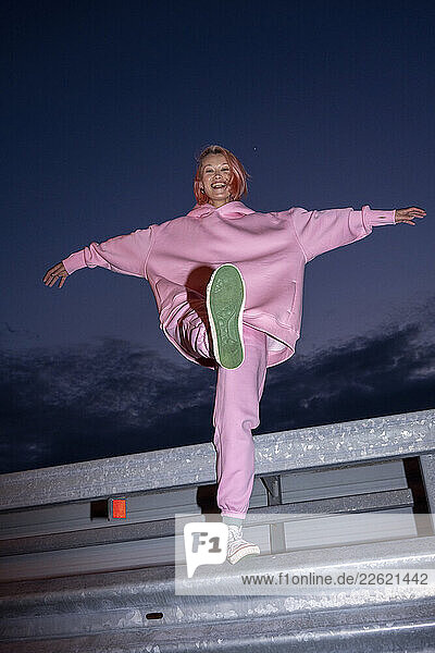 Young woman with pink hair wearing pink track suit standing on one leg on road barrier at night